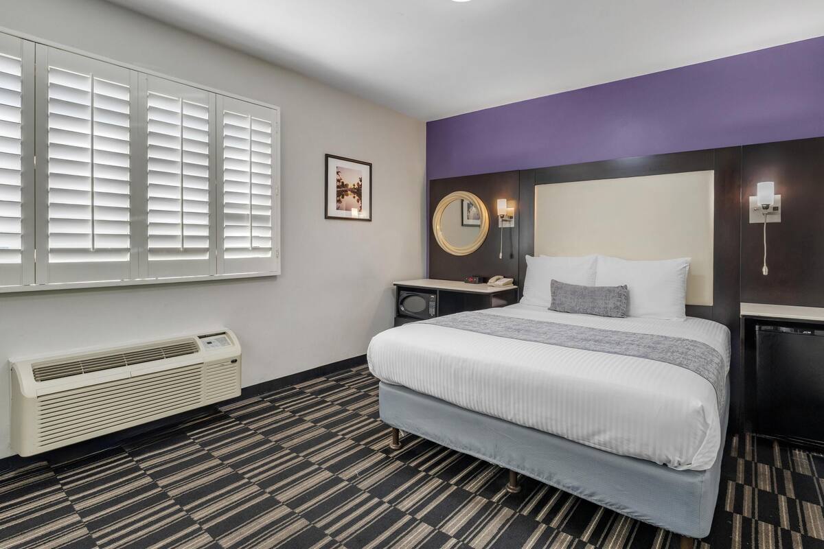 A hotel room with a bed, a bedside table, a mirror, a wall-mounted phone, and a window with blinds.