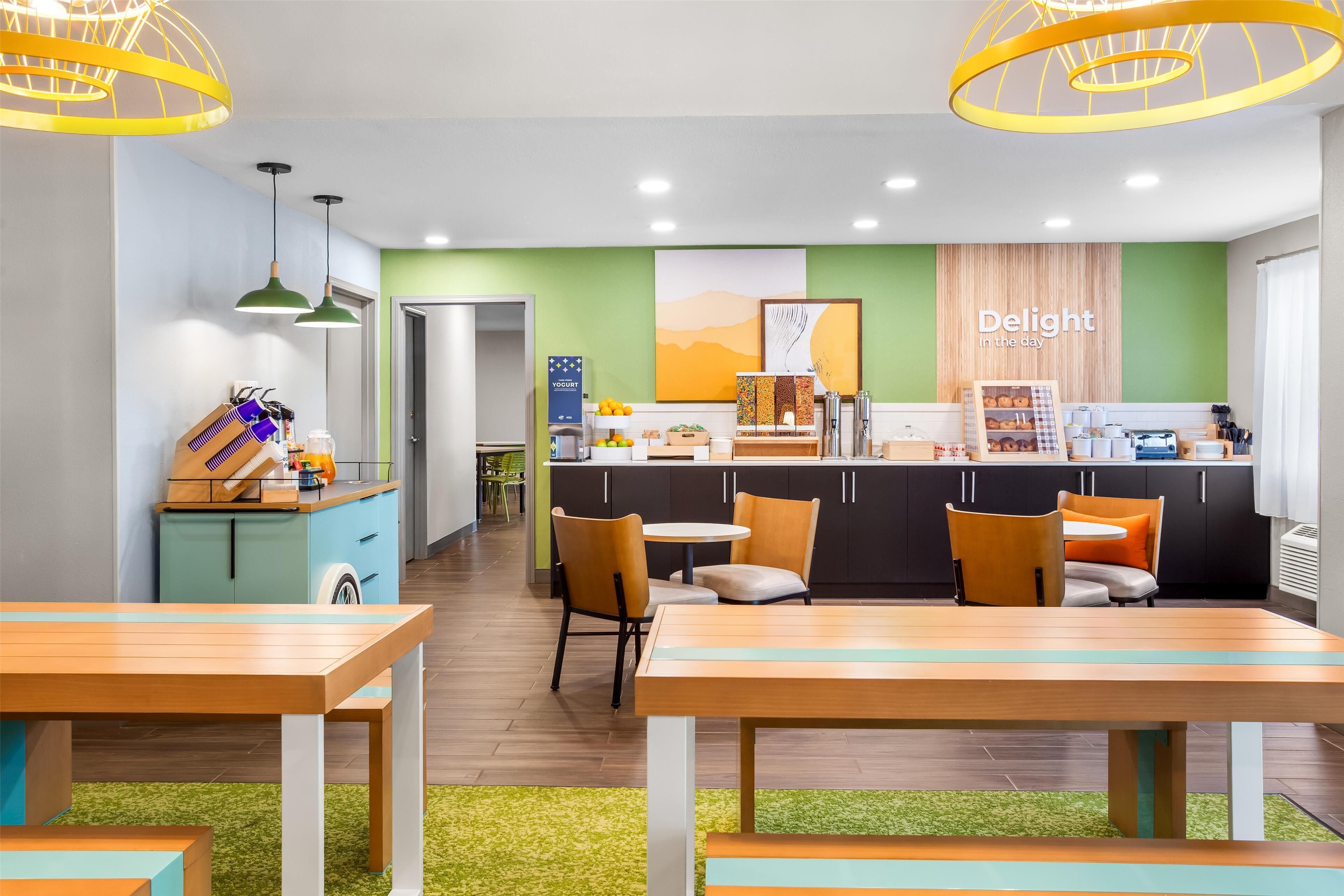 Une cuisine moderne dotée d’un îlot central, d’un coin repas avec des tables en bois et d’un mur d’accent vert orné d’une inscription : « Savourez chaque instant ».
