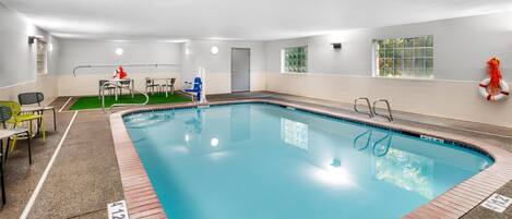 An indoor swimming pool with a clear blue water, surrounded by a tiled deck. There are chairs and tables on the left side, and a lifebuoy on the right wall.