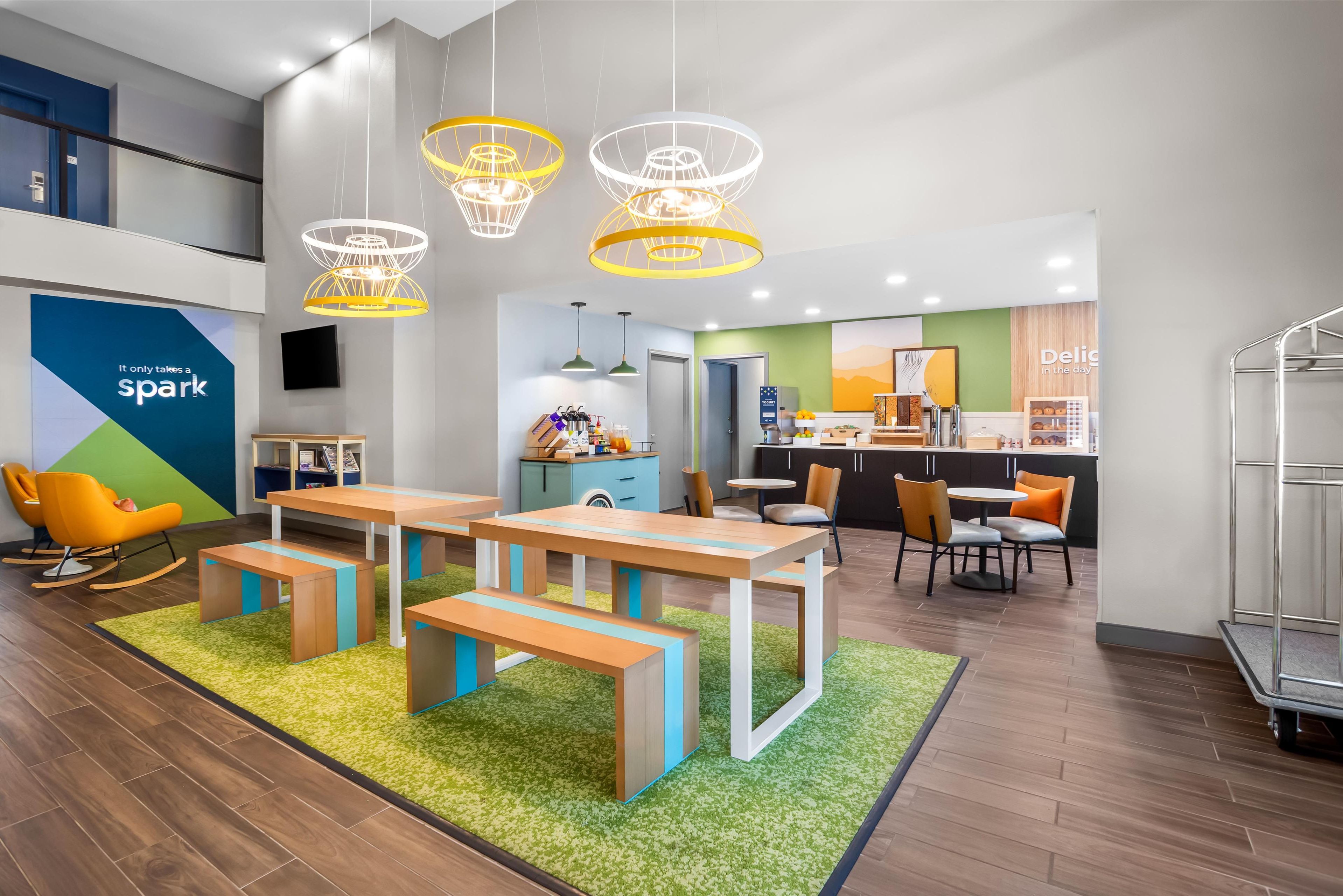 A modern dining area with wooden tables and benches, a kitchen in the background, and a sign that reads "It only takes a spark."