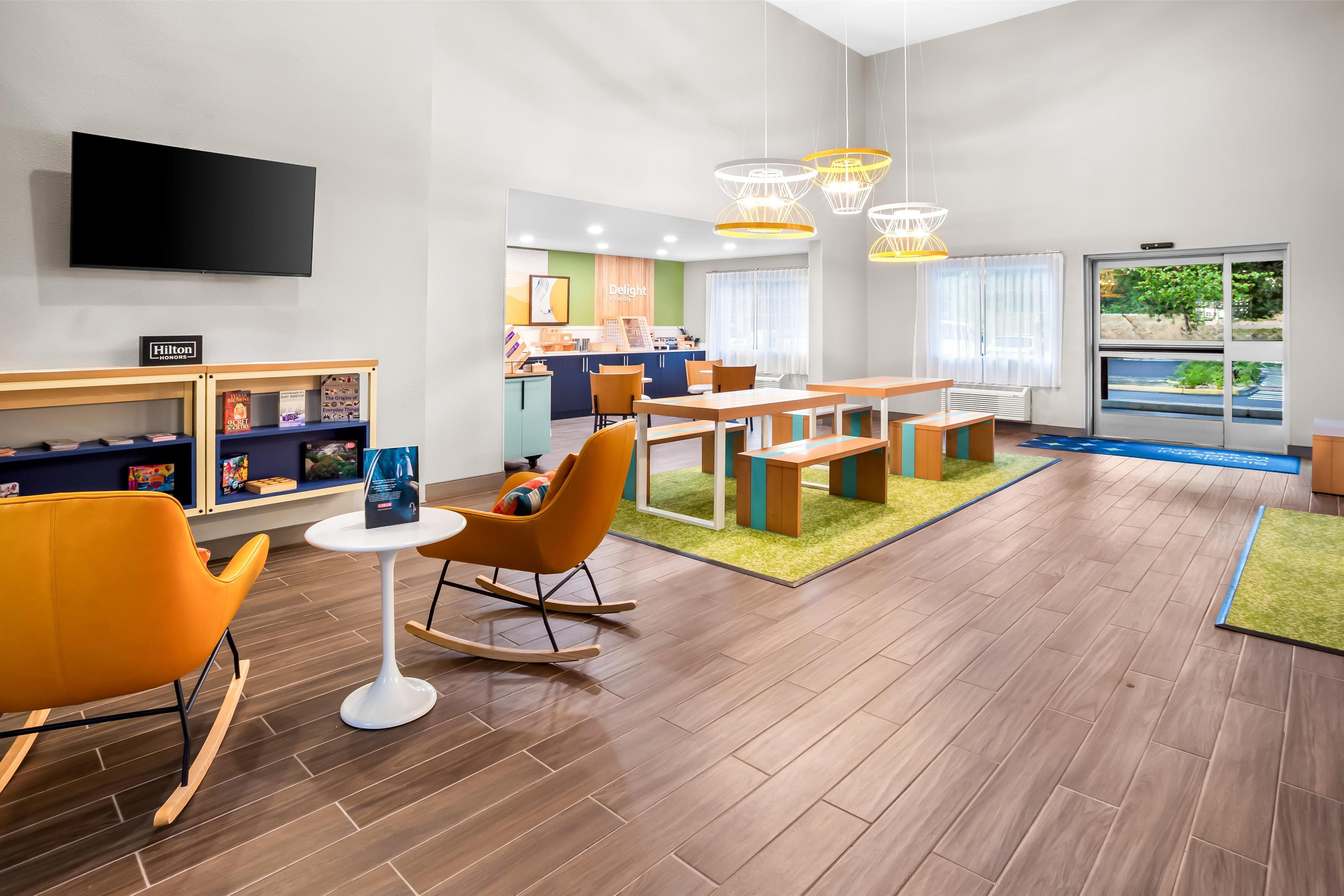 A modern hotel lobby with a flat-screen TV, a children's play area, a dining area with wooden furniture, and a large window offering a view of the outdoors.
