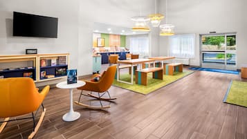 A modern hotel lobby with a flat-screen TV, a children's play area, a dining area with wooden furniture, and a large window offering a view of the outdoors.