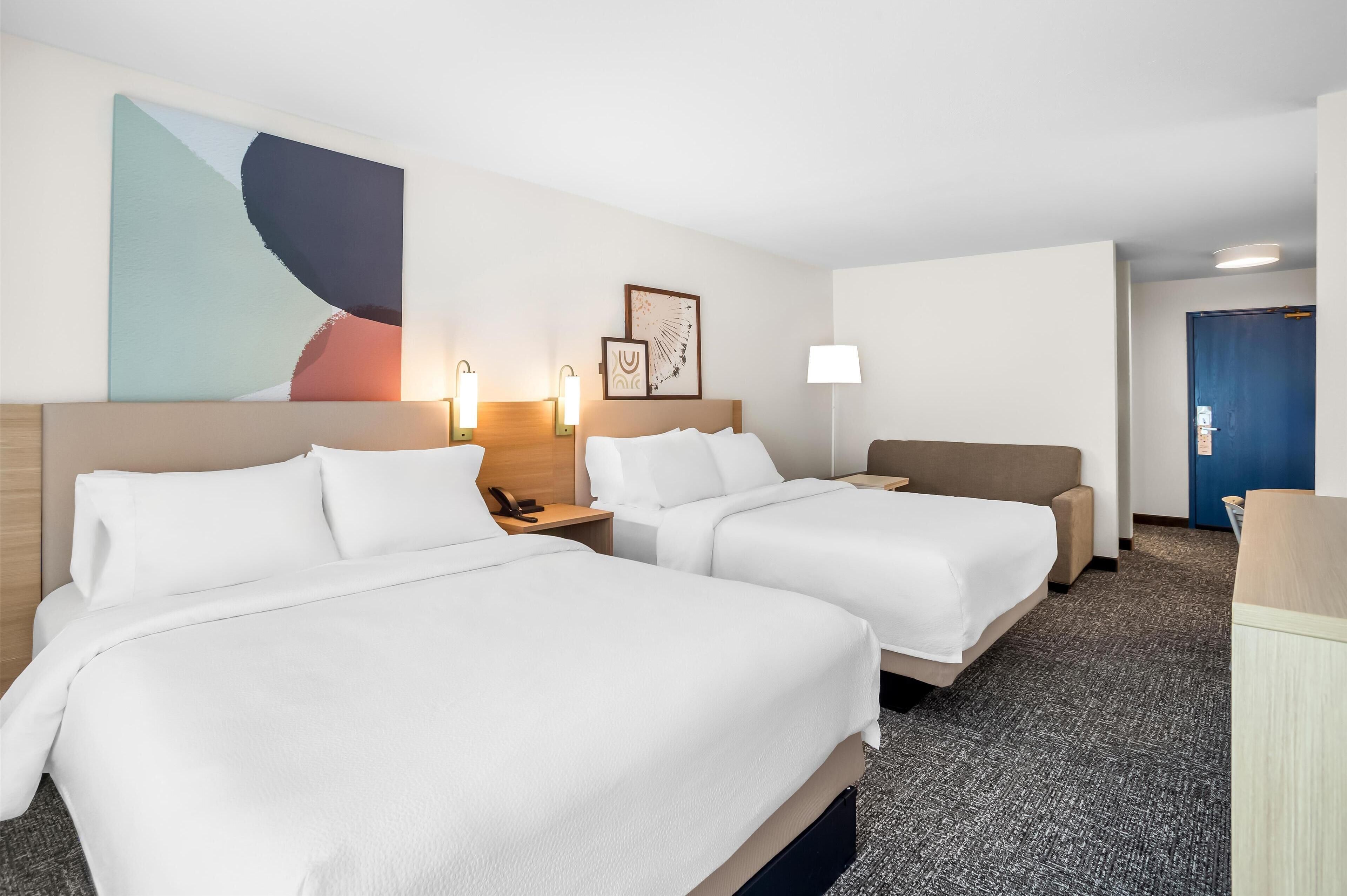 Een hotelkamer met twee bedden, een bureau, een stoel, een lamp en kunst aan de muur.