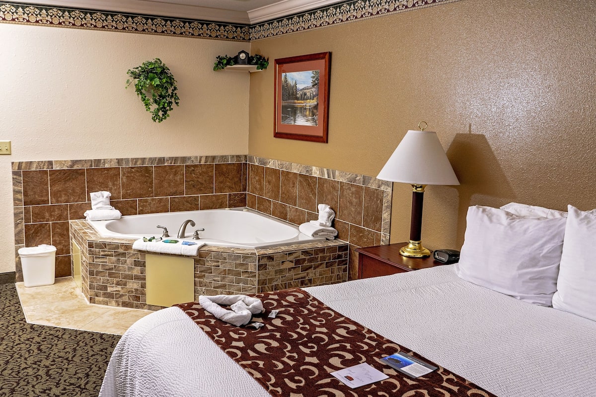 Quarto de hotel com uma cama grande, um banheiro com banheira, uma mesinha de cabeceira com abajur e um quadro pendurado na parede.