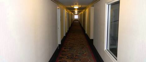 Un pasillo largo y estrecho de hotel con alfombra estampada, paredes blancas y una lámpara en el techo.