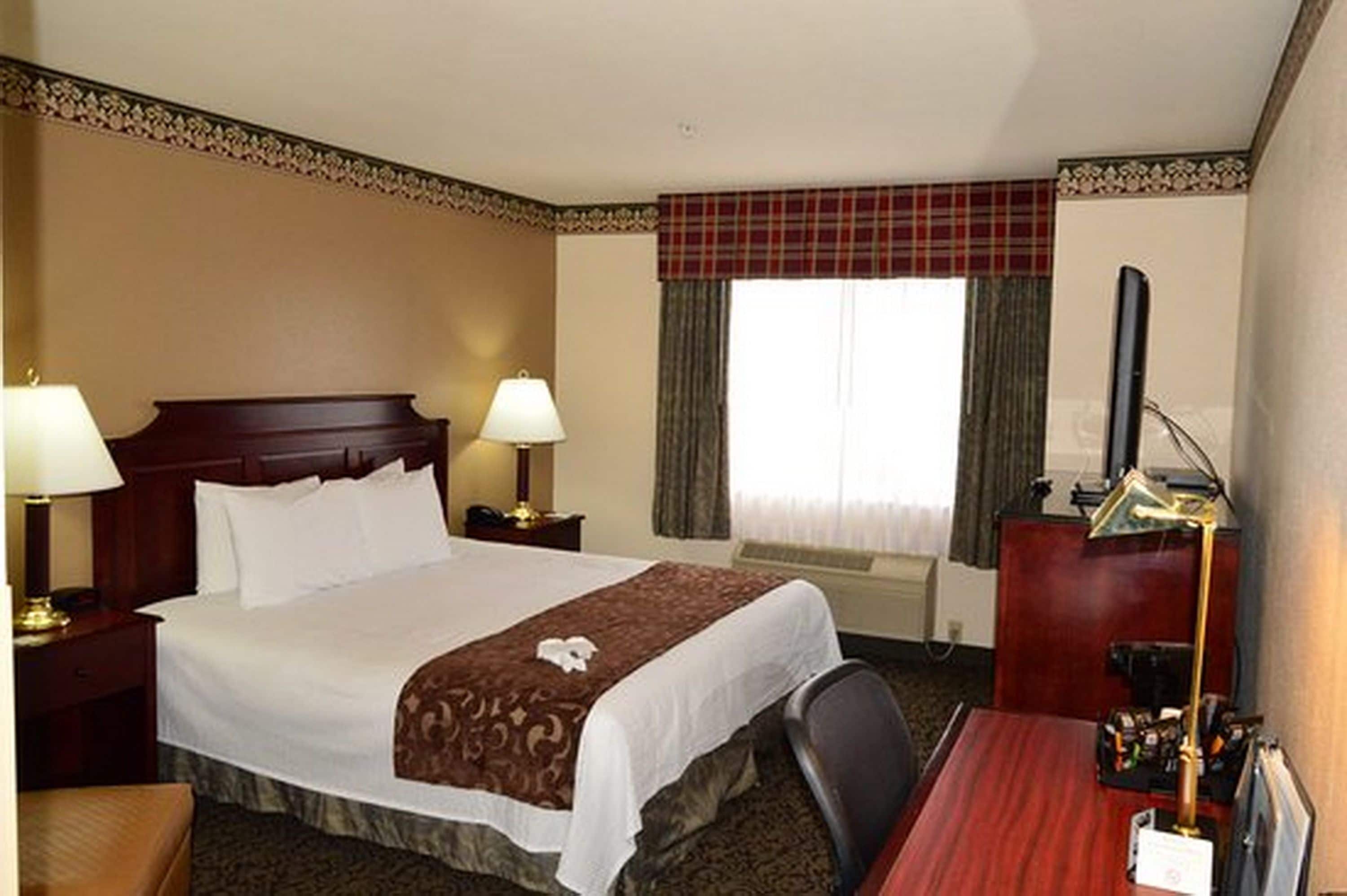 A hotel room with a large bed, two bedside tables with lamps, a desk with a chair, a TV, and a window with curtains.
