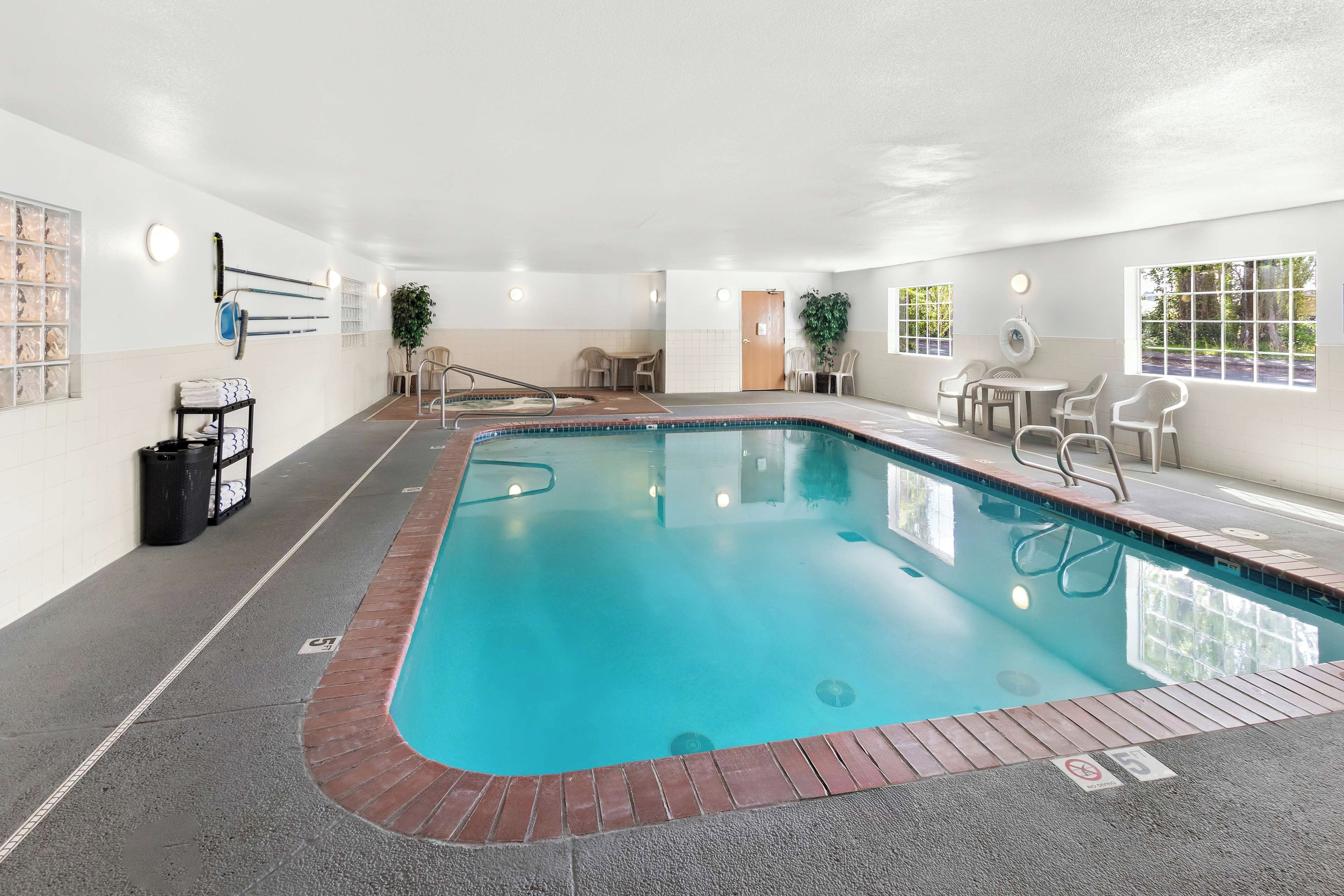 An indoor swimming pool with a clear blue water, surrounded by a tiled floor and a brick border. There are chairs and tables around the pool, and a door leading to another room.