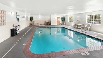 An indoor swimming pool with a clear blue water, surrounded by a tiled floor and a brick border. There are chairs and tables around the pool, and a door leading to another room.