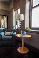 Een moderne hotelkamer met een donker leren bank, een ronde houten tafel met een lamp, en een raam met uitzicht op de stad.