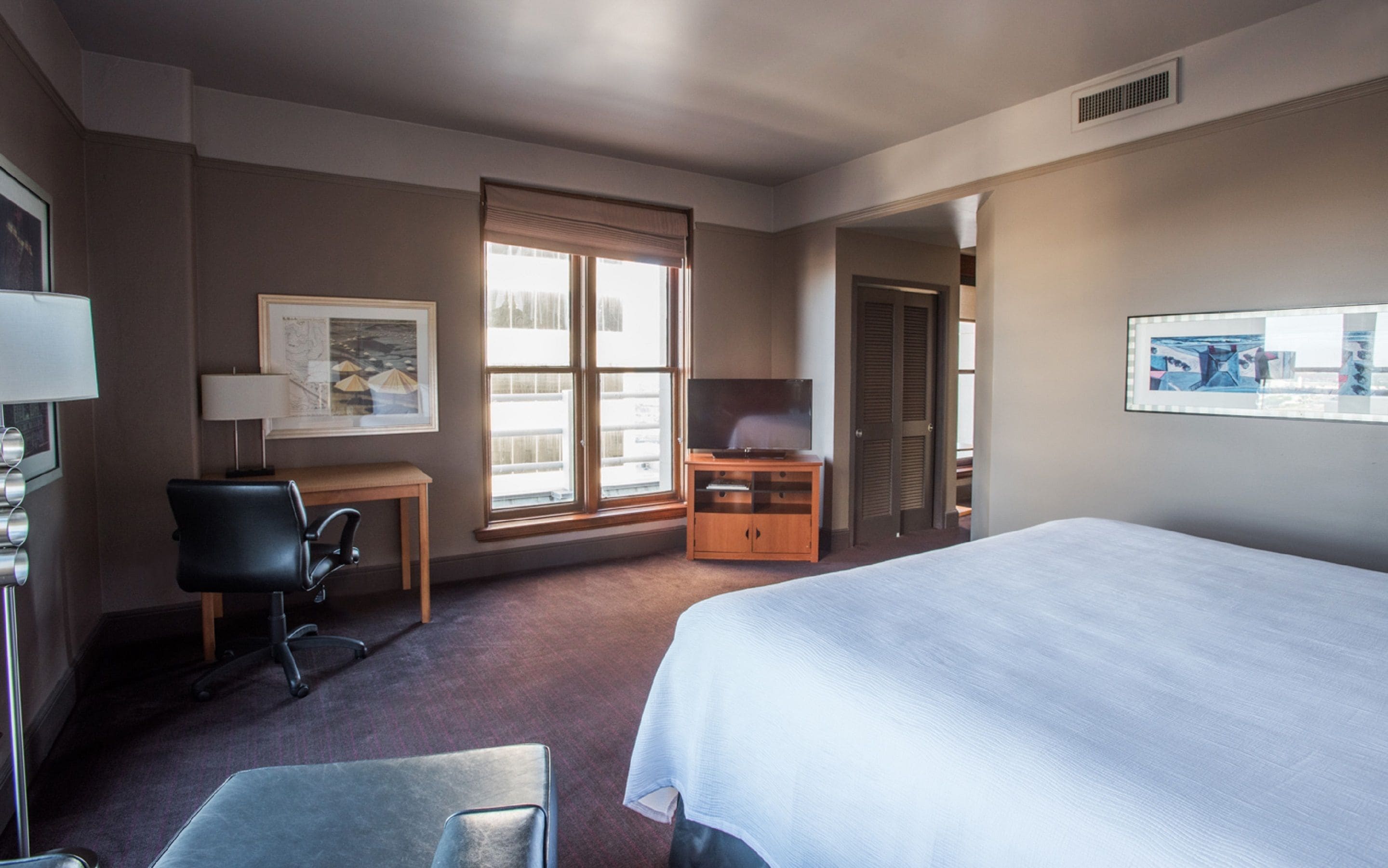 A hotel room with a bed, a desk with a chair, a television, and a window with blinds.