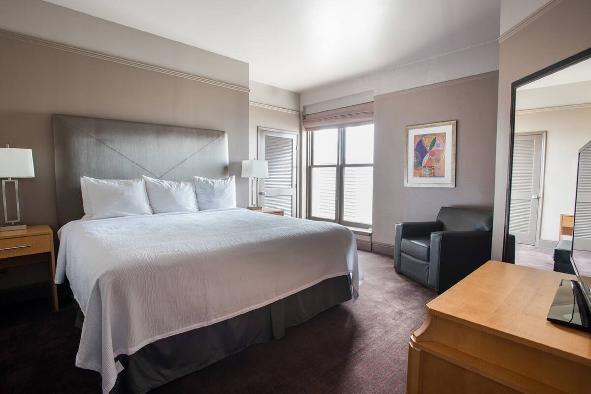 A hotel room with a large bed, a desk, a chair, and a window with blinds.