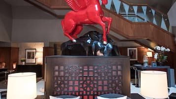 Un moderno vestíbulo de hotel con un área de estar central que presenta un sofá negro con cojines blancos, una pared decorativa con patrones circulares y una escultura de un caballo rojo sobre un pedestal negro.