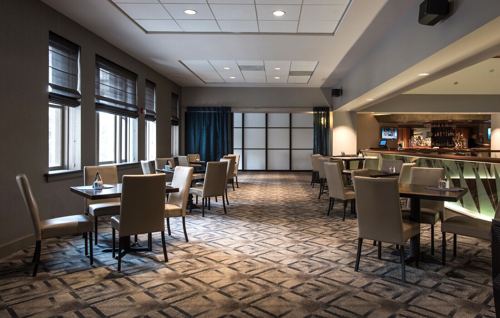 A hotel dining area with multiple tables and chairs, a bar area, and large windows with blinds.