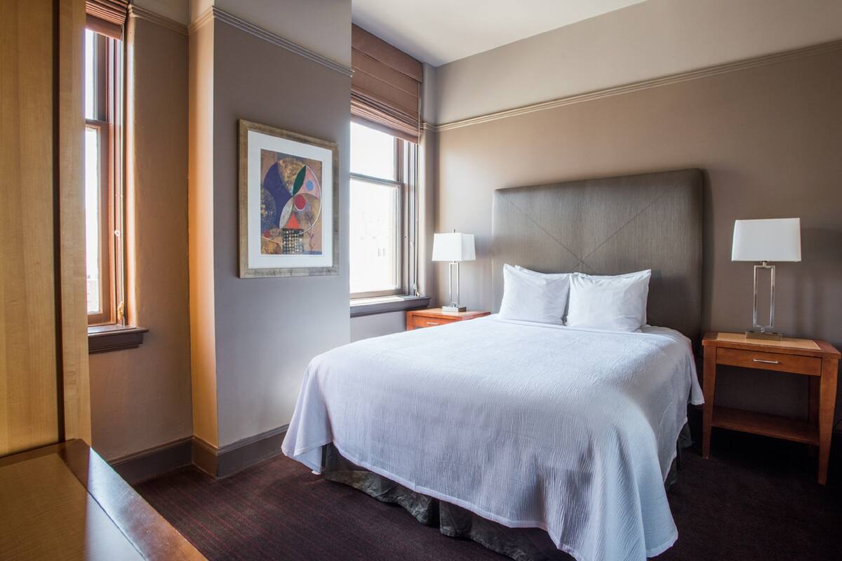 A hotel room with a large bed, two bedside tables with lamps, a window with blinds, and a framed artwork on the wall.