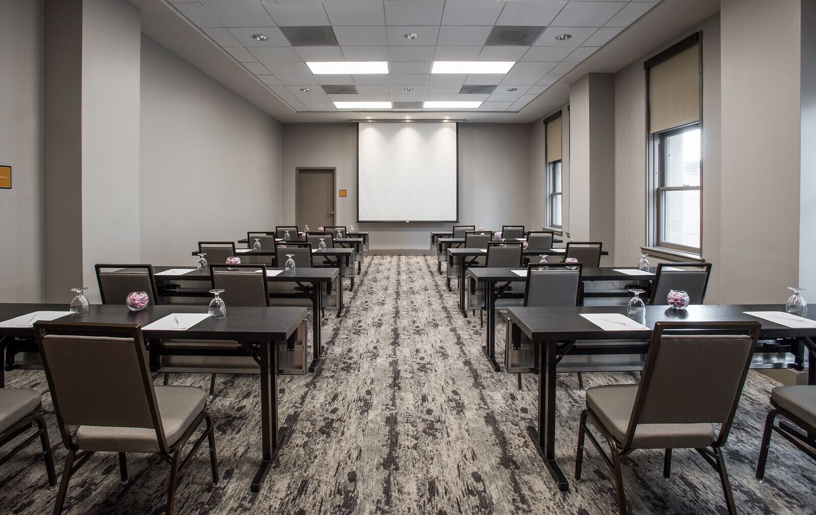A conference room with rows of tables and chairs, a projector screen, and a carpeted floor.