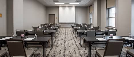 A conference room with rows of tables and chairs, a projector screen, and a carpeted floor.