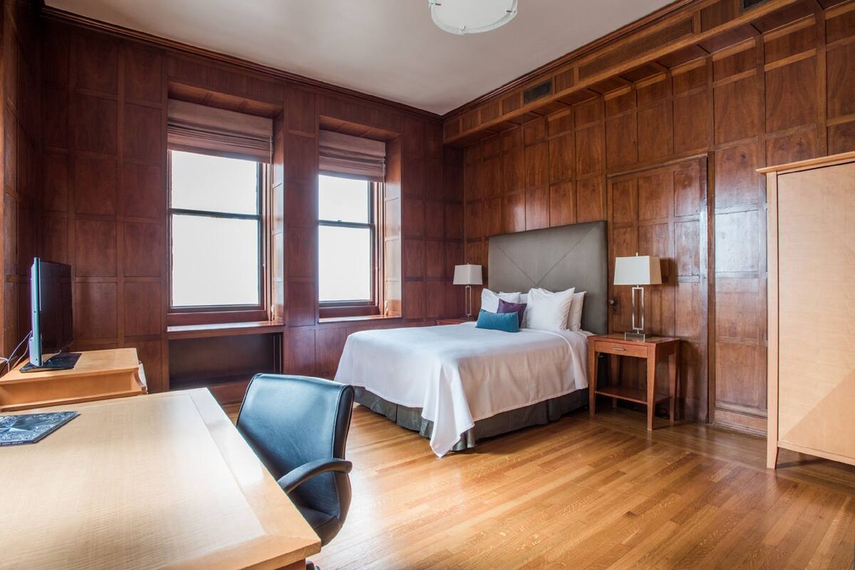 A hotel room with a wooden paneled wall, a bed with white bedding, two bedside tables with lamps, a desk with a chair, and a television on a stand.