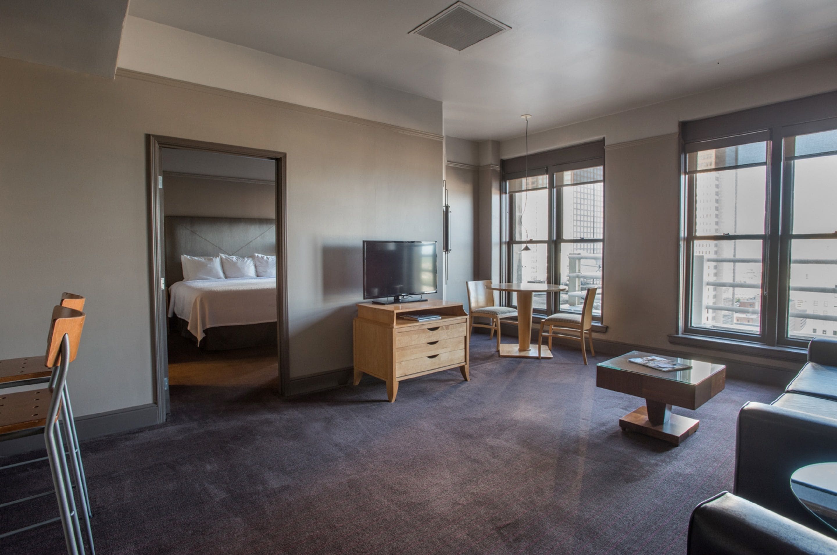 Une chambre d’hôtel moderne équipée d’un lit, d’une télévision, d’un bureau et de grandes fenêtres donnant sur la ville.