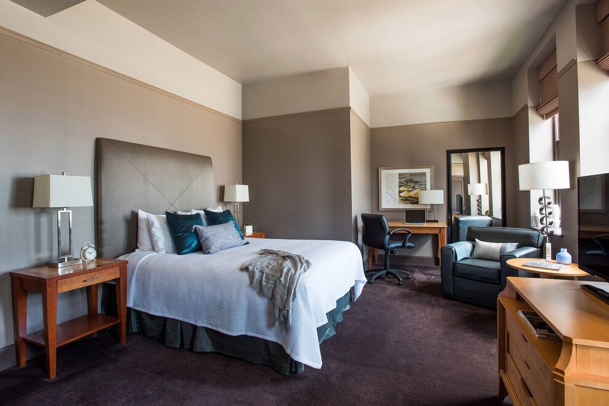 A hotel room with a large bed, a desk, a chair, a TV, and a lamp.