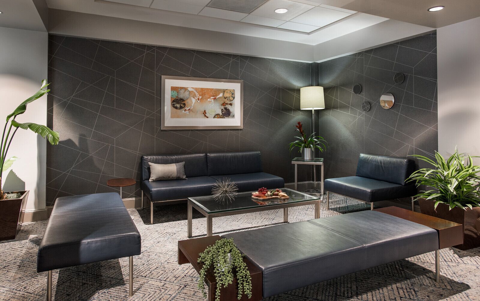 Un salon moderne avec un mur à motifs géométriques de couleur sombre, une table basse centrale, deux canapés, une lampe sur pied et des plantes en pot.