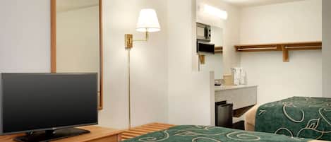 Habitación de hotel con televisor sobre un atril de madera, dos camas con cubrecamas de estampado verde, un espejo montado en la pared, una lámpara y una pequeña zona de kitchenette.