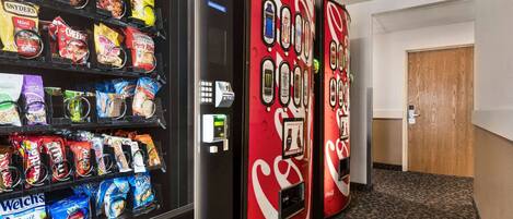 Una máquina expendedora con snacks y bebidas, una máquina con la marca Coca-Cola, y una puerta de madera al fondo.