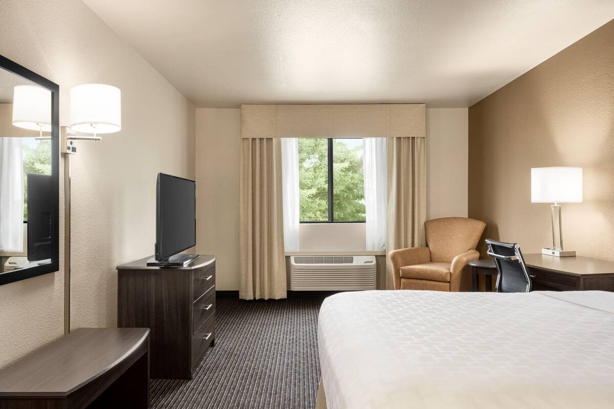 Una habitación de hotel con cama, escritorio, silla, televisor y ventana con cortinas.