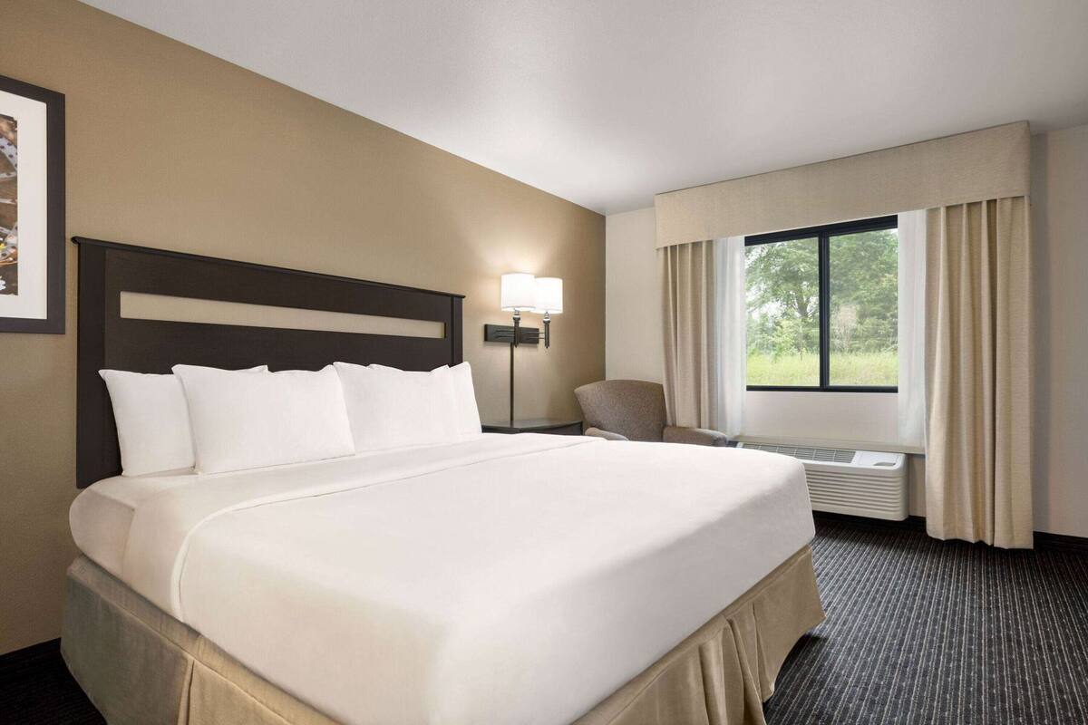 Habitaci ón de hotel con una cama grande, una silla, una mesita de noche con lámpara y una ventana con cortinas.