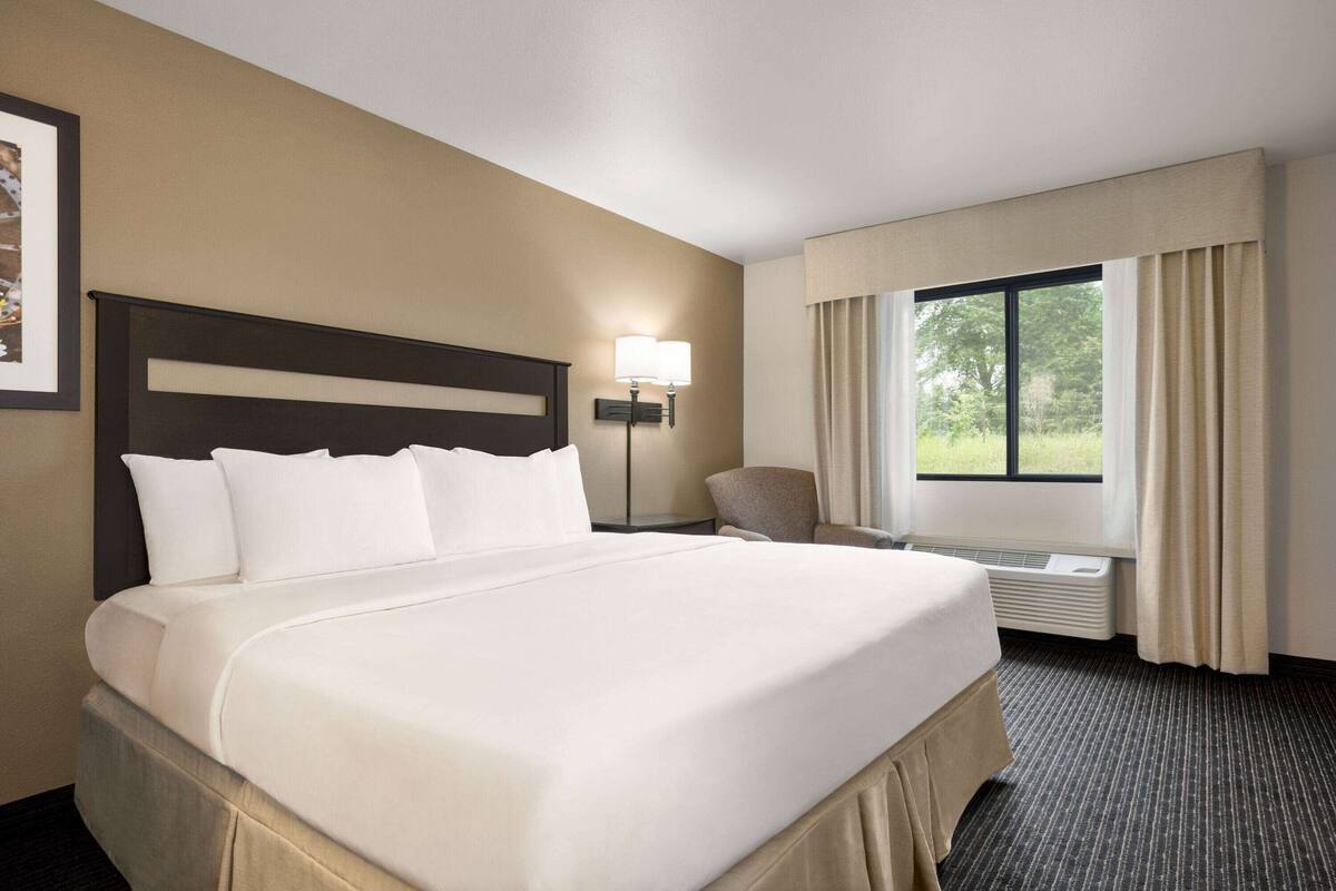 Habitación de hotel con una cama grande, una silla, una mesita de noche, una lámpara y un ventanal con cortinas.