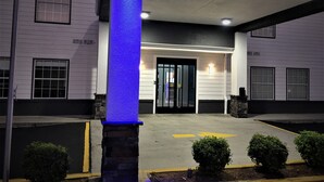 Un edificio con un pilar iluminado en azul, un acceso cubierto y un estacionamiento con flechas direccionales.