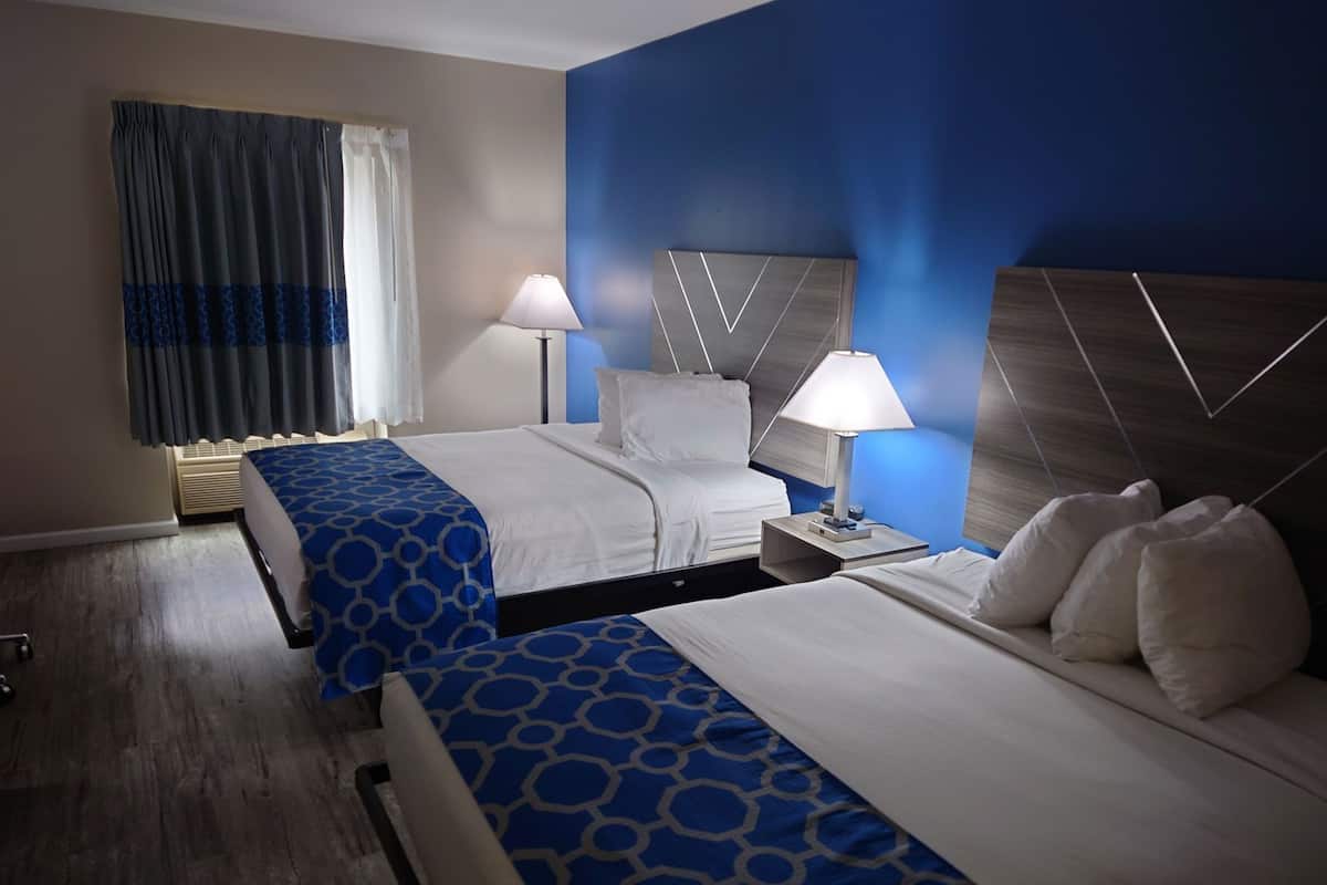 Habitación de hotel con dos camas, una mesita de noche, una lámpara, una ventana con cortinas y una pared con diseño geométrico.