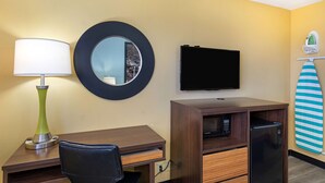 A hotel room with a desk, chair, a flat-screen TV, a mirror, a refrigerator, and a surfboard on the wall.