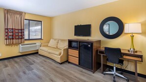 A hotel room with a sofa, a desk with a chair, a television, a mirror, and a lamp.