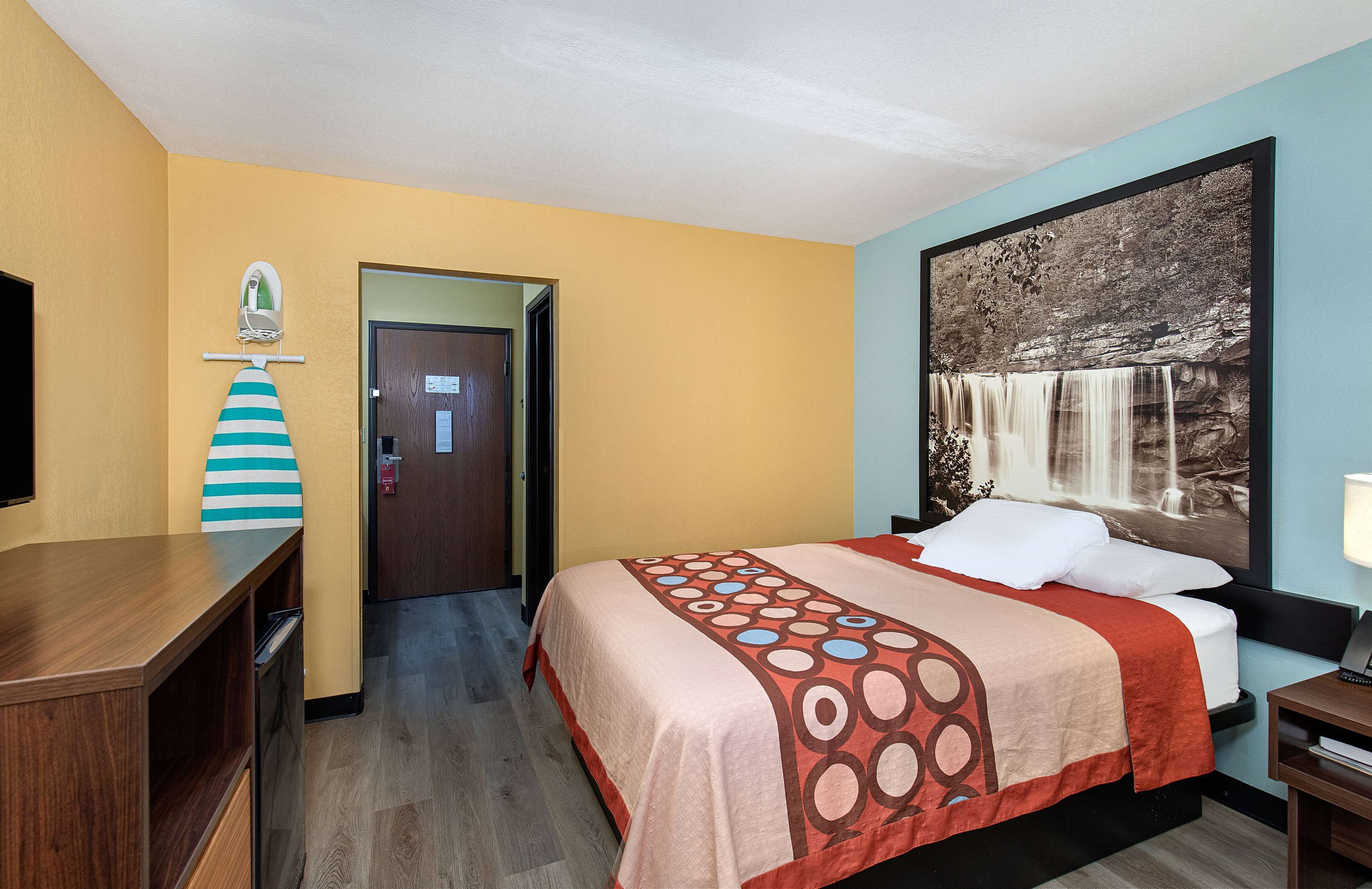A hotel room with a bed, a desk, a chair, a door, and a wall with a waterfall painting.