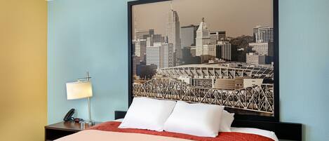 A hotel room with a bed, a nightstand, a lamp, and a large cityscape painting on the wall.