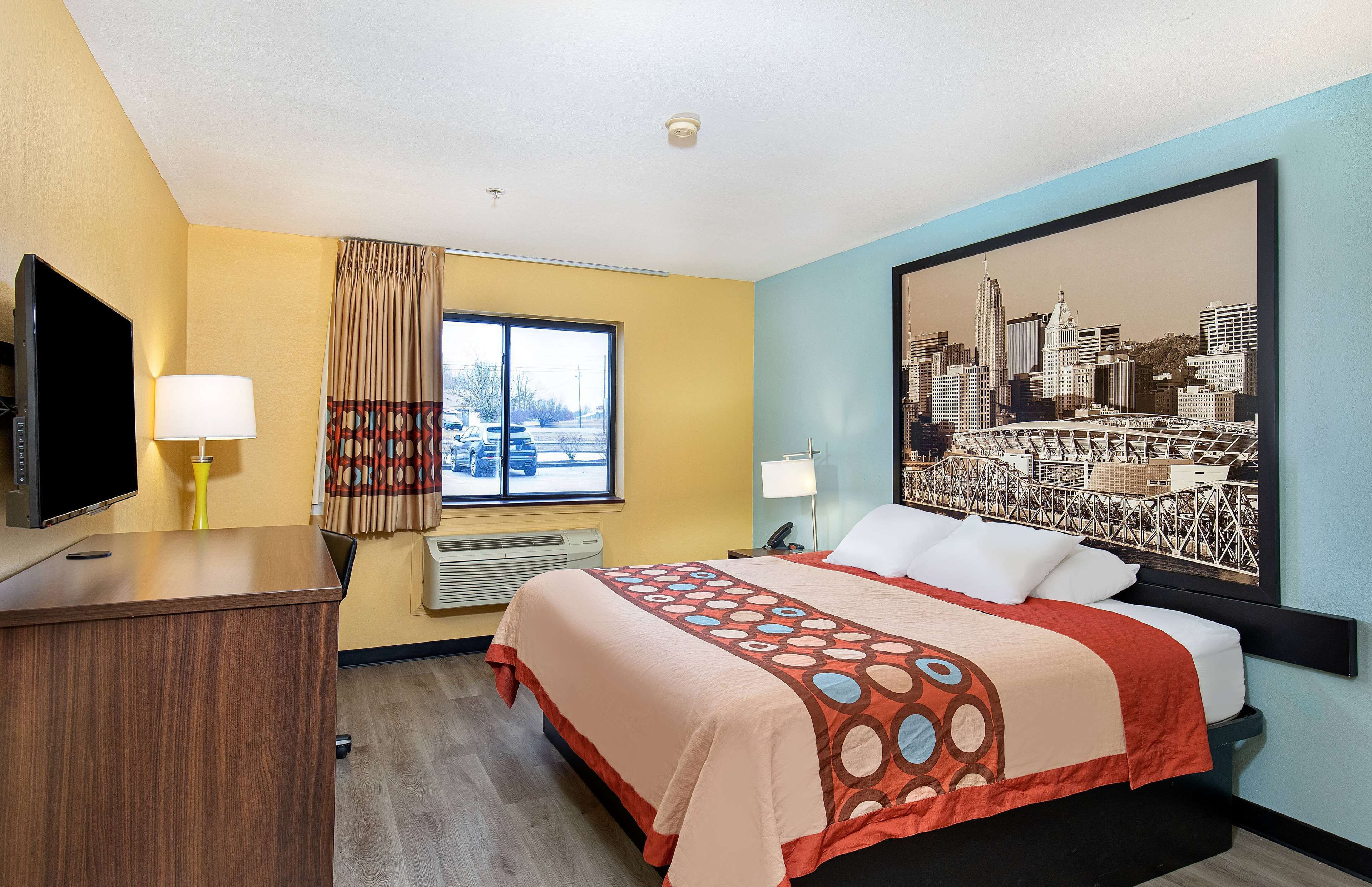 A hotel room with a bed, a desk with a TV, a window with curtains, and a cityscape painting on the wall.