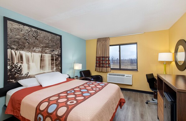 A hotel room with a large bed, a desk, a chair, a window with curtains, and a wall-mounted mirror.