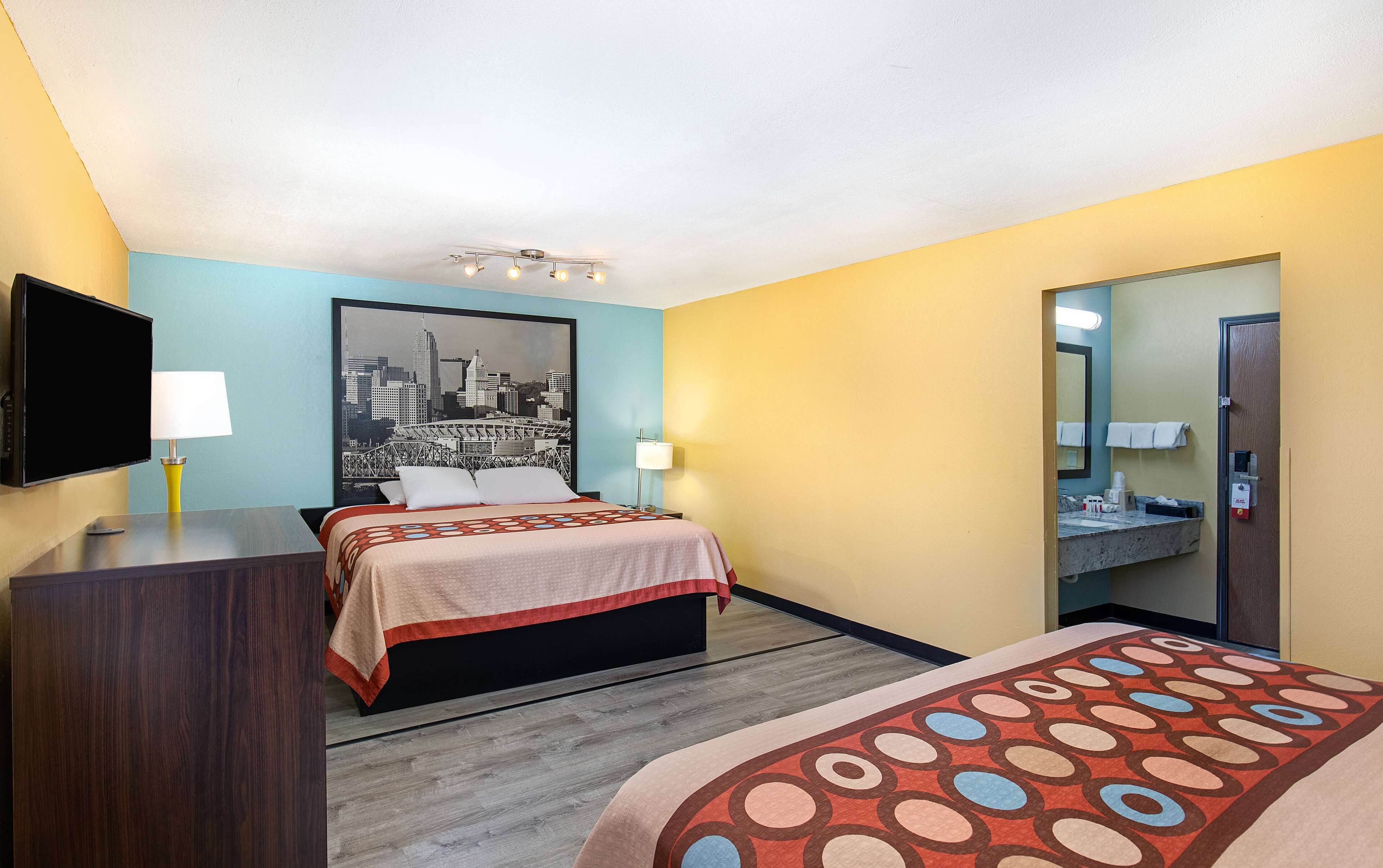 A hotel room with a bed, a desk, a television, a cityscape painting, and a bathroom mirror.