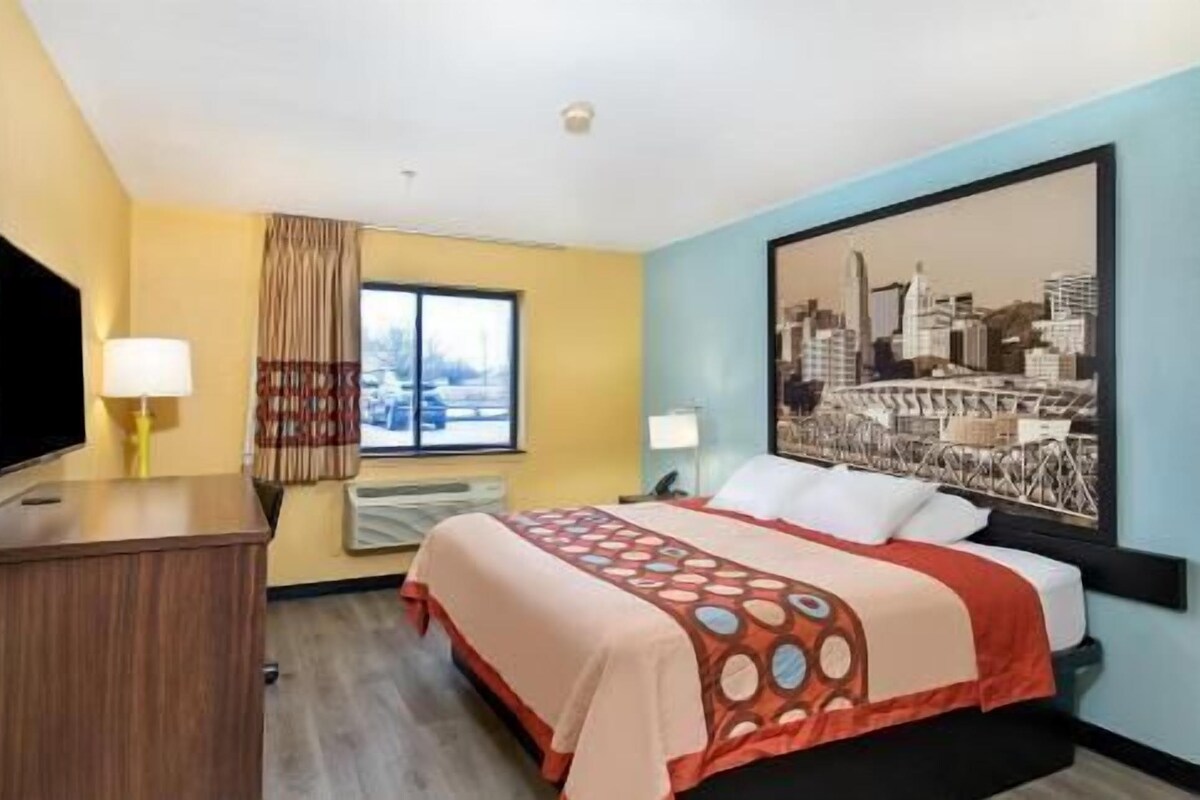 Una habitación de hotel con cama, escritorio, televisor, ventana con cortinas y un cuadro de paisaje urbano en la pared.