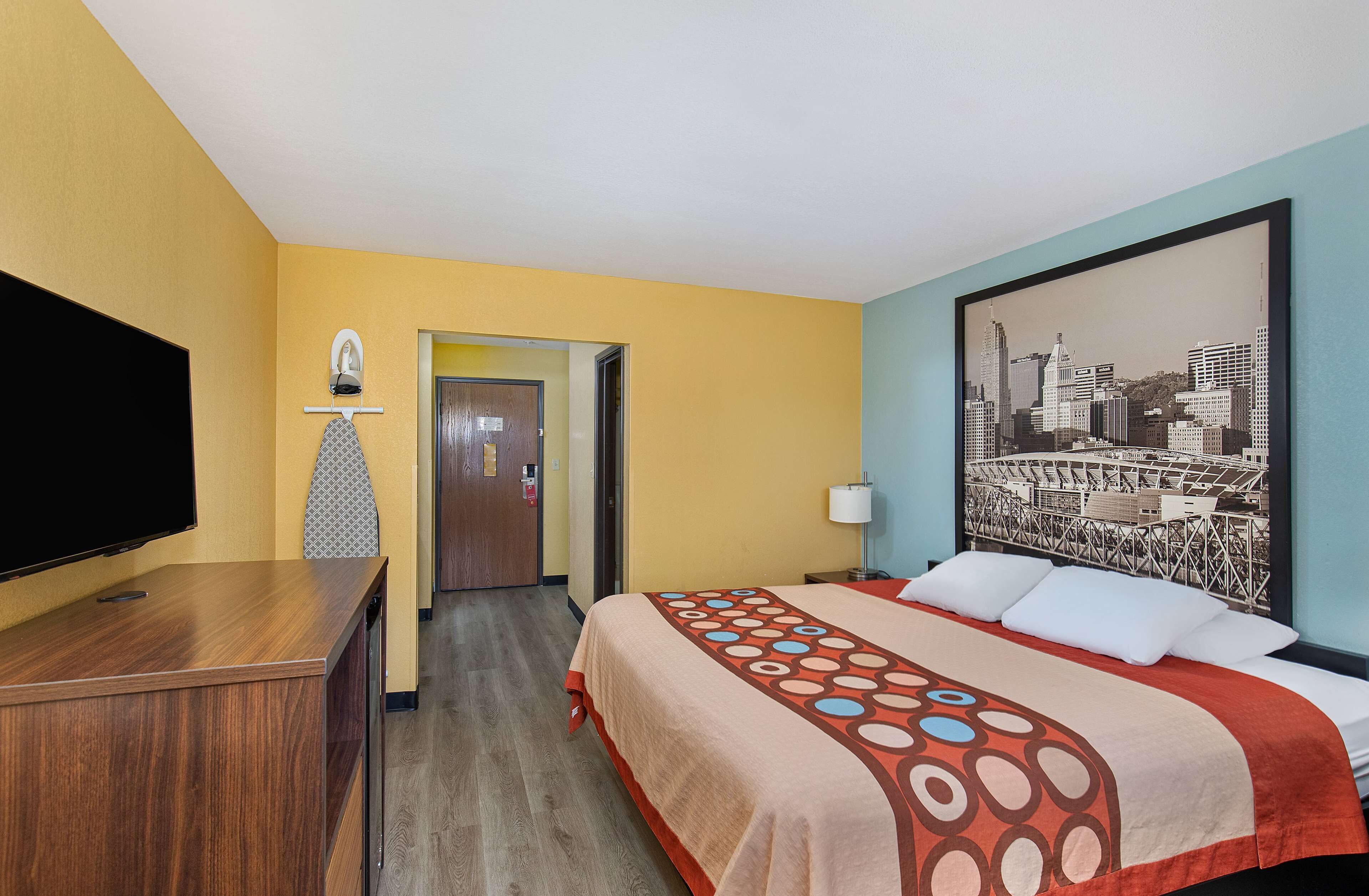 A hotel room with a bed, a television, a desk, and a cityscape painting on the wall.