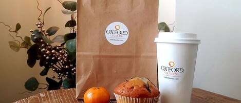 A brown paper bag and a white cup with the Oxford Suites logo, a muffin, and an orange on a wooden table.