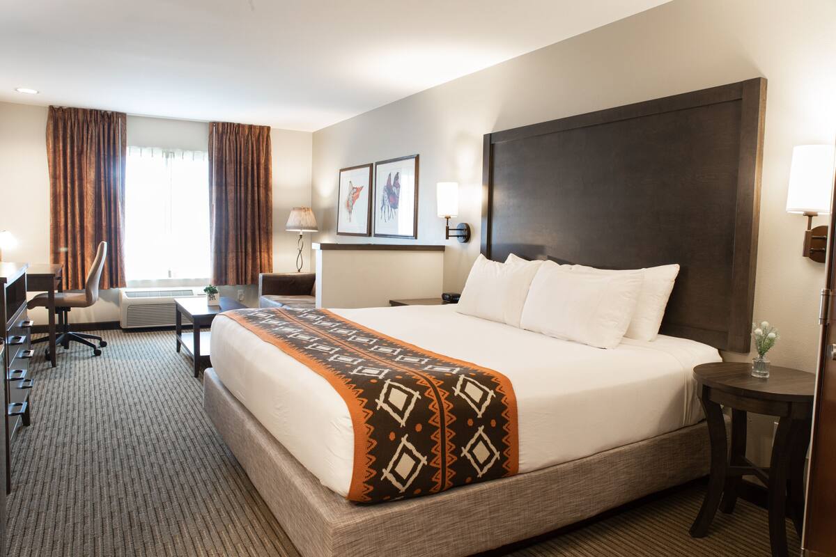 A hotel room with a large bed, a desk, a chair, a lamp, and a window with curtains.