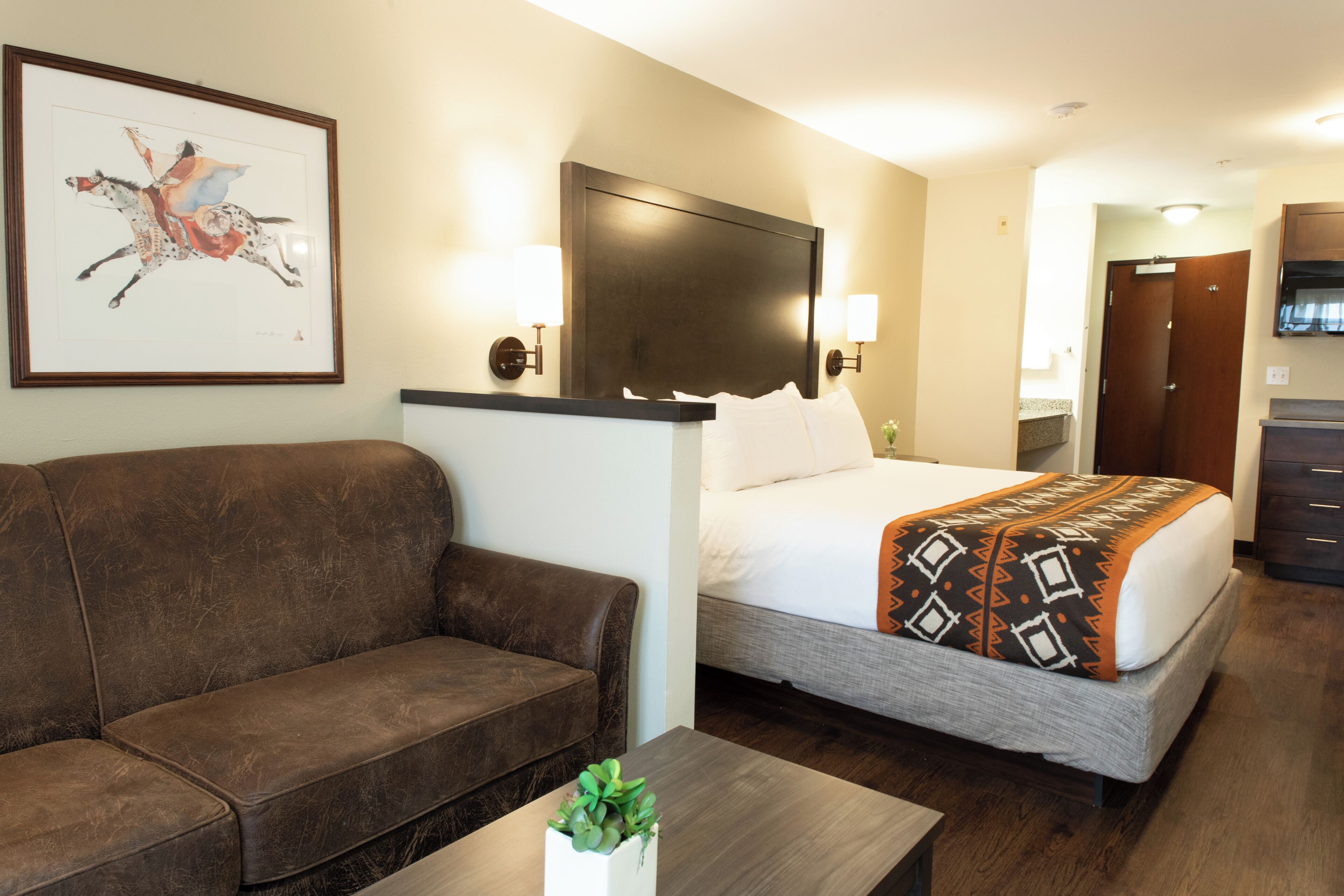 Quarto de hotel com uma cama, um sofá, uma mesinha de cabeceira com uma planta e um quadro pendurado na parede.