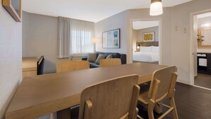 A modern hotel room with a wooden dining table, two chairs, a sofa, and a large window with curtains.