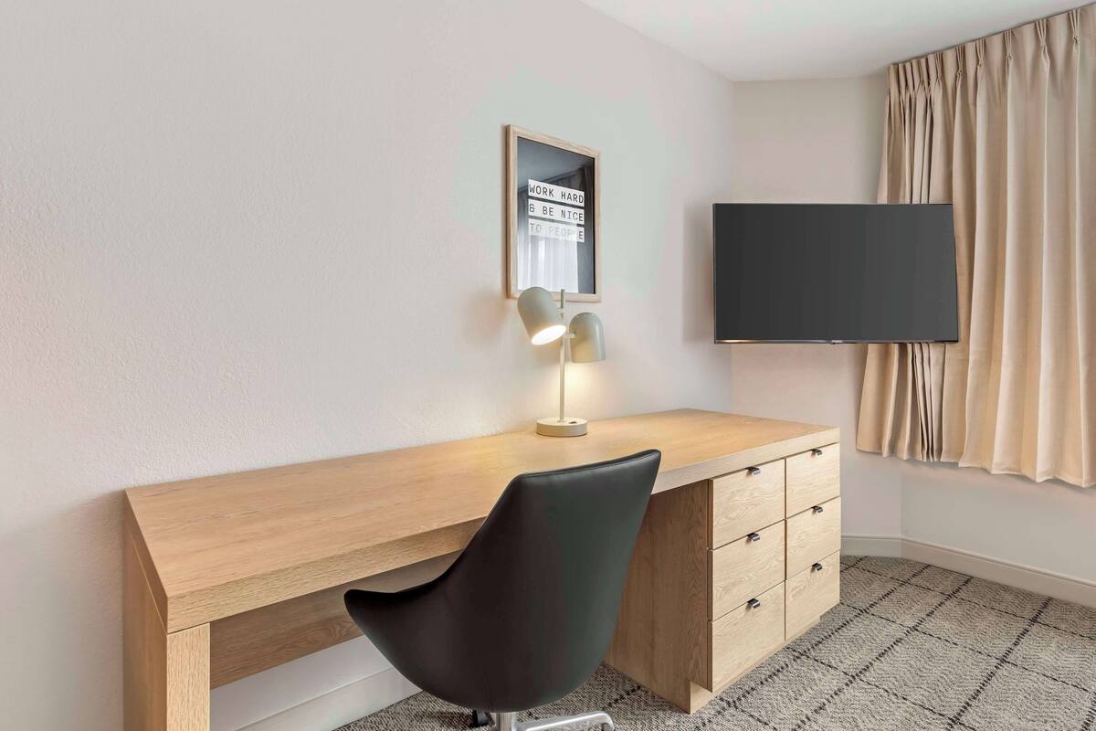 Een moderne hotelkamer met een houten bureau, een zwarte bureaustoel, een bureaulamp, een wandtelevisie en een grote spiegel.