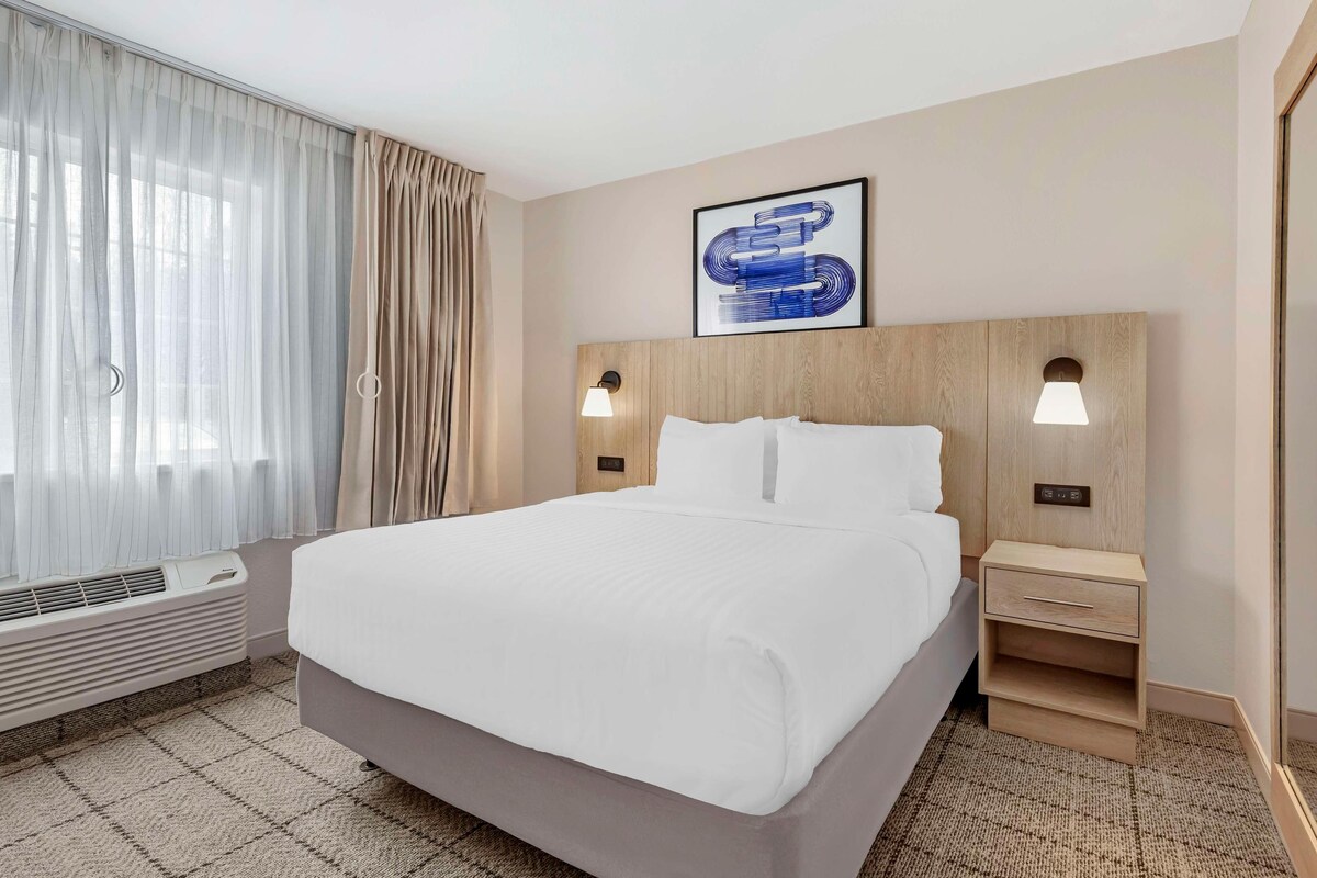 Une chambre d’hôtel avec un grand lit, deux tables de chevet, une lampe fixée au mur, un tableau encadré et une fenêtre avec des rideaux.