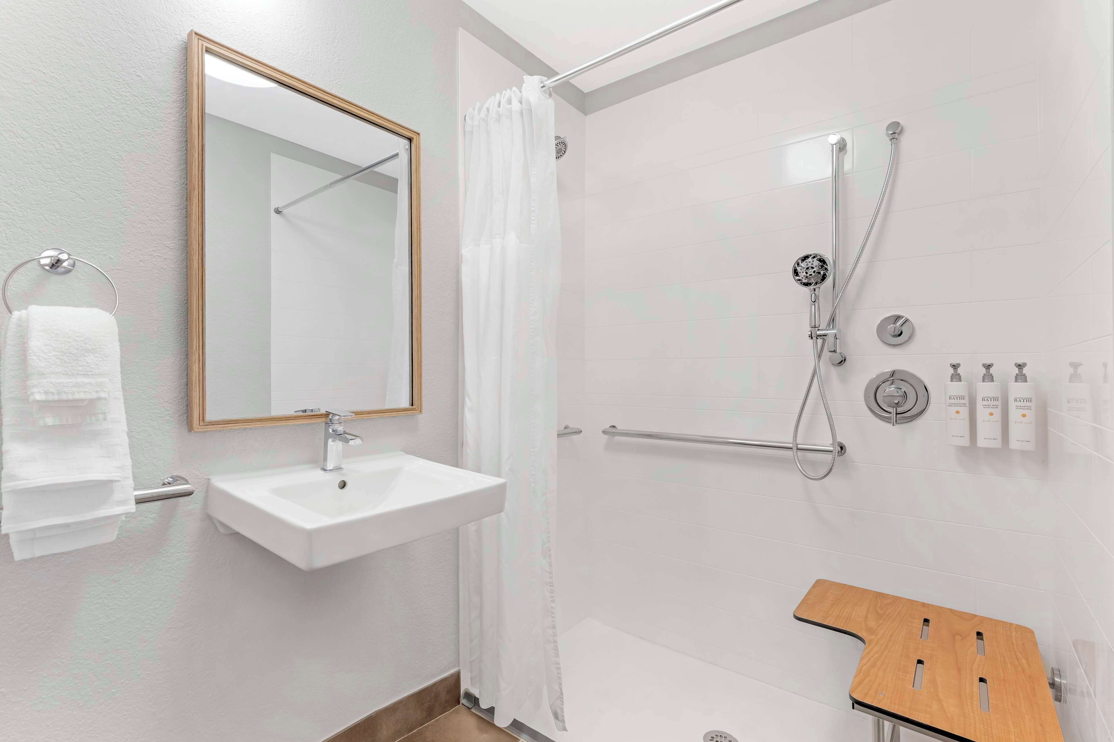 Un baño moderno con un lavamanos blanco, un espejo, una ducha con cabezal de ducha manual y un taburete de madera.