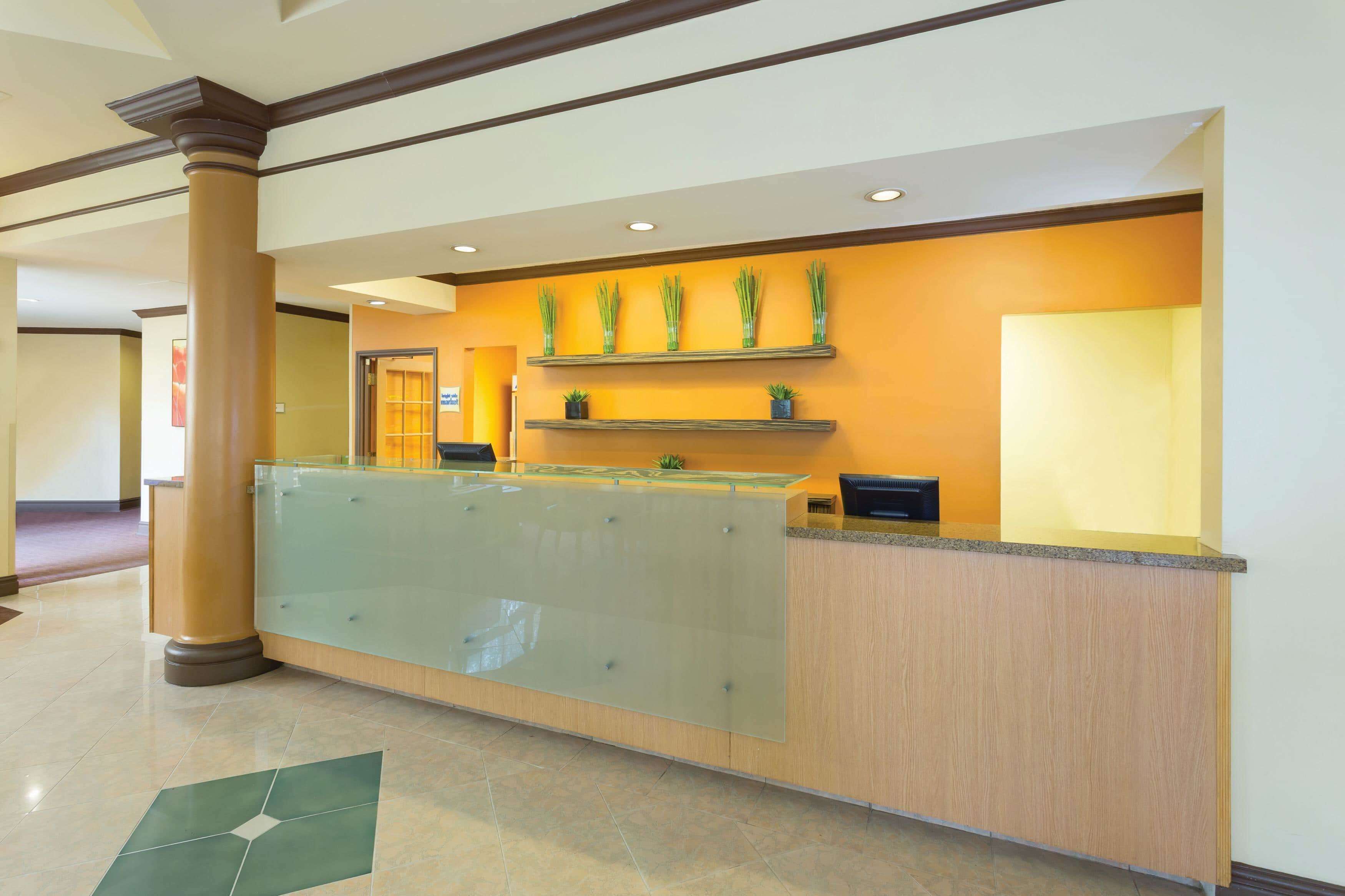 Un área de recepción con un separador de vidrio, mostrador de madera y plantas decorativas en estanterías.