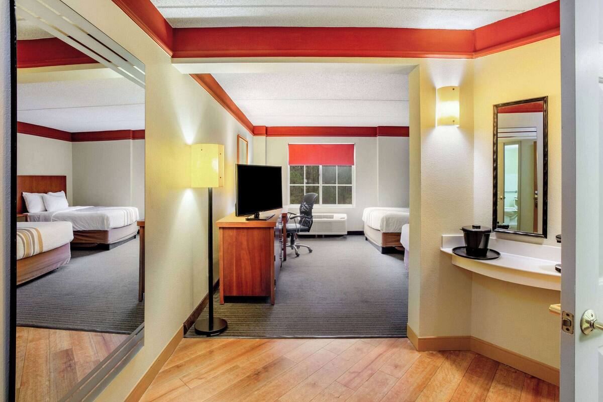 Habitación de hotel con dos camas, un escritorio, una silla, un televisor, una ventana y un baño con lavamanos y espejo.