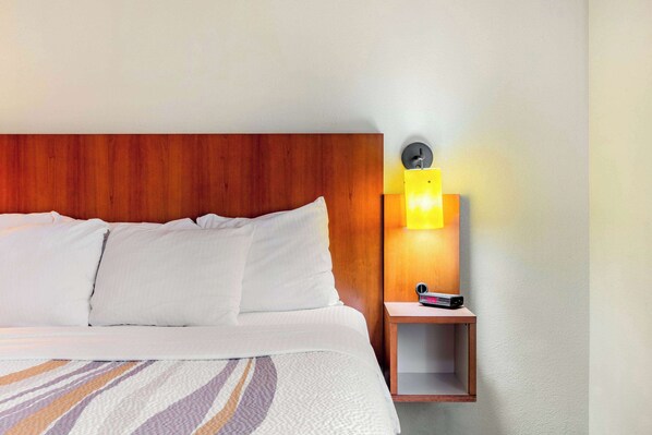 A neatly made bed with white linens and a patterned bedspread, a wooden headboard, and a bedside table with a lamp and a clock.